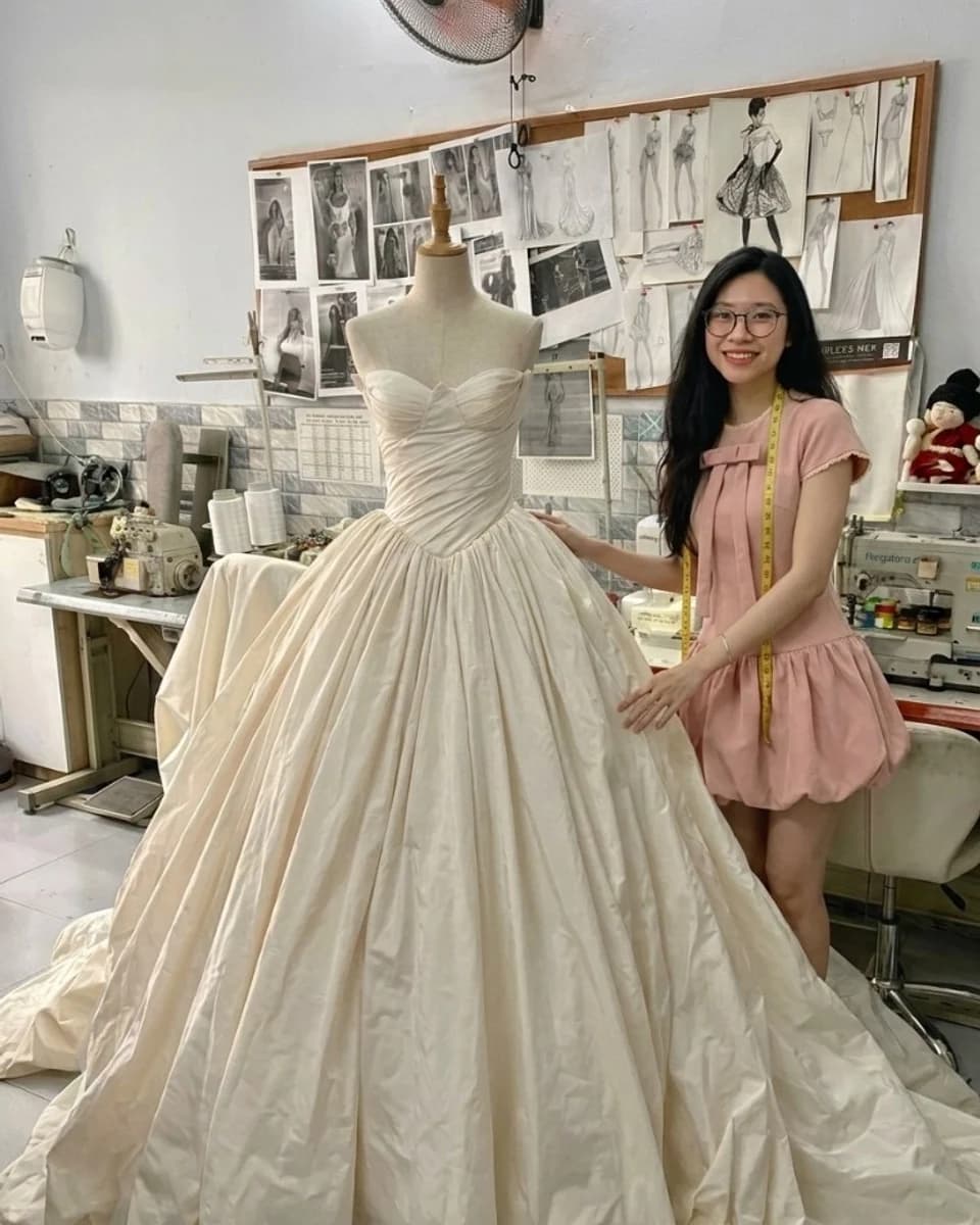 Vi standing beside a wedding gown on a dress form in the VI DRESSMAKERS working space